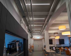 Blick in eine moderne Kantine mit linearer Deckenbeleuchtung und runden Pendelleuchten über Sitzbereichen.
