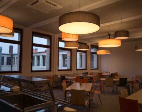 Blick in eine moderne Kantine mit linearer Deckenbeleuchtung und runden Pendelleuchten über Sitzbereichen.