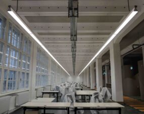 Lange LED-Leuchten verlaufen parallel über einer Reihe von Arbeitsplätzen in einem großen, lichtdurchfluteten Raum.