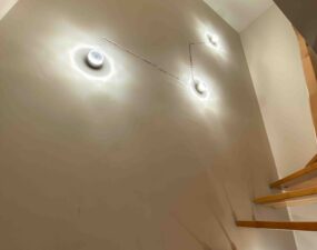 Runde Wandlampen noch ohne Verkleidung beleuchten die Wand entlang einer Holztreppe.