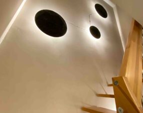 Runde Wandlampen mit indirektem Licht beleuchten die Wand entlang einer Holztreppe.