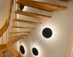 Runde Wandlampen mit indirektem Licht beleuchten die Wand entlang einer Holztreppe.