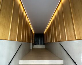 Beleuchteter Eingangsbereich mit Treppe zum Aufzug in der Französischen Straße in Berlin, mit warmem, indirektem Licht, das entlang der Holzverkleidung an den Wänden verläuft.