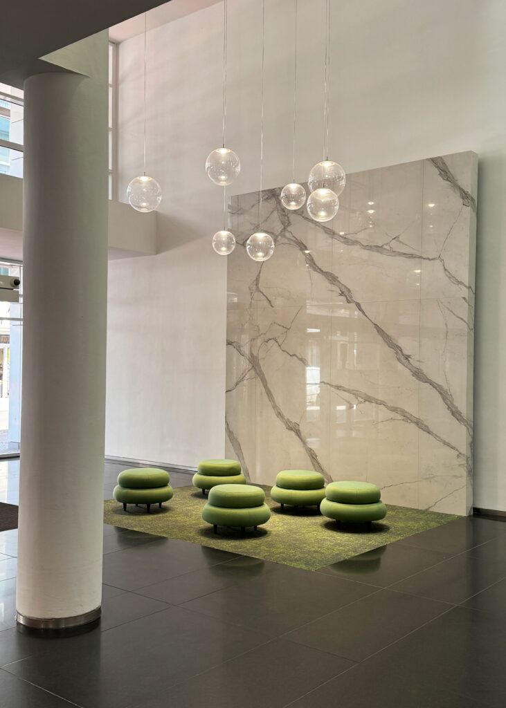 Beleuchtung in der Empfangshalle der Friedrichstraße 55 in Berlin, mit schwebenden, runden Glaskugellampen. Im Vordergrund ist eine Sitzgelegenheit mit grünen, runden Poufs auf einem grünen Teppich.