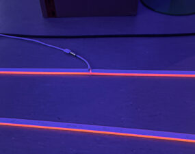Zwei parallel verlegte, orange-violett leuchtende LED-Streifen auf dem Boden mit sichtbarer Kabelverbindung.