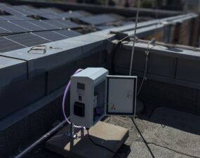 Geöffnetes weißes Schaltgehäuse mit violett leuchtendem LED-Streifen auf einem Flachdach, im Hintergrund Solarpaneele und Berliner Dächer.