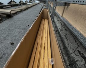 Blick von oben in einen offenen, langgestreckten Karton mit gebündeltem LED-Profilmaterial, verlegt entlang einer Dachattika, im Hintergrund Solarpaneele und Gebäudeaufbauten.