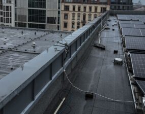 Nasses Flachdach mit verlegten Kabeln entlang einer Attika, im Hintergrund Berliner Altbaufassaden bei bewölktem Himmel.